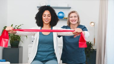 Kendini adamış bir kıdemli fizyoterapist direniş bandı egzersizleri yapan genç bir siyahi müşteriye uzman rehberliği sağlıyor. Bu terapi seansı, küçük bir uygulama ortamında kişisel bakım ve etkili rehabilitasyonu vurgular.. 