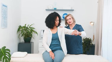 Şefkatli bir kıdemli fizyoterapist parlak, modern bir fizyoterapi kliniğinde genç bir siyahi hastaya detaylı bir kol ve omuz muayenesi yapar.. 