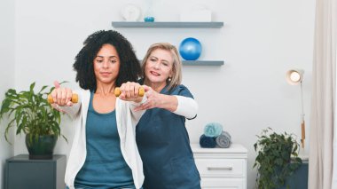 Deneyimli bir kıdemli fizyoterapist, muhtemelen bir işyeri sahibi, genç bir Black müşterisine, temiz, organize edilmiş küçük muayenehanesinde silah rehabilitasyonu için çeşitli hareket alıştırmalarında dikkatle rehberlik eder.. 