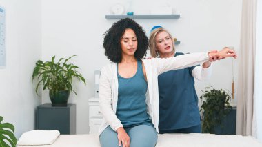 Yetenekli bir kıdemli fizyoterapistin genç siyahi müşterilere omuz ve kol tedavisi uygulamasını izleyin. Bu sahnede profesyonel spor fizyoterapisi ve rehabilitasyon hizmetleri küçük bir alanda sunulmaktadır.