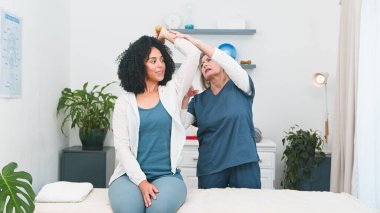 Genç bir Black müşterisi, temiz ve modern bir klinikte kıdemli bir fizyoterapistten kişiselleştirilmiş fizyoterapi görüyor. Uzman, ona kol ve omuz hareketlerinde rehberlik ediyor, rehabilitasyon için gerekli bakım ve desteği sağlıyor.