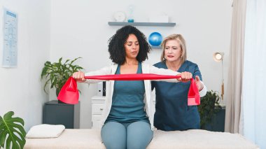 Kendini adamış bir kıdemli fizyoterapist direniş bandı egzersizleri yapan genç bir siyahi müşteriye uzman rehberliği sağlıyor. Bu terapi seansı, küçük bir uygulama ortamında kişisel bakım ve etkili rehabilitasyonu vurgular.. 