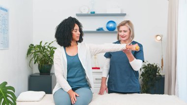 Üst düzey bir fizyoterapist, genç siyahi bir müşteriye kol egzersizinde yardımcı olur. Küçük, profesyonel bir uygulamada düzgün bir form gösterir. Daha iyi sağlık ve spor sağlığını destekleyen bu rehabilitasyon oturumu. 