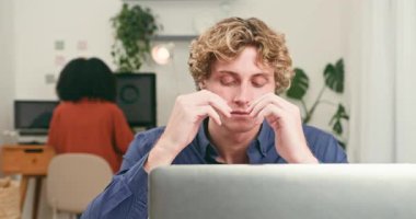 Genç bir adam, gözle görülür bir şekilde stresli, bilgisayarının üzerinde çalışırken şakaklarını ovalıyor çağdaş bir teknoloji şirketinde. Arka planda çalışan farklı bir meslektaşım, yoğun ve odaklanmış bir çevreyi vurguluyor..