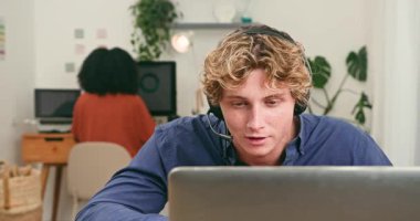 Kulaklık takan neşeli genç bir adam laptopuyla meşgul oluyor, muhtemelen canlı ve modern bir teknoloji firmasının ofisindeki bir video görüşmesinde. Çeşitli bir meslektaş özenle arka planda çalışarak, işbirlikçi ve yenilikçi bir çalışma ortamını ön plana çıkarıyor.