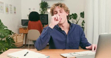 Genç bir adam, modern bir teknoloji firmasında özenle çalışırken yorulduğunu göstererek esniyor. Çeşitli meslektaşları arka planda çalışarak, çağdaş çalışma ortamlarını ve üretkenliği vurguluyor..