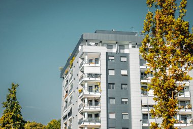 Mavi gökyüzü ve yeşil ağaçları olan modern bir binanın ön cephesi