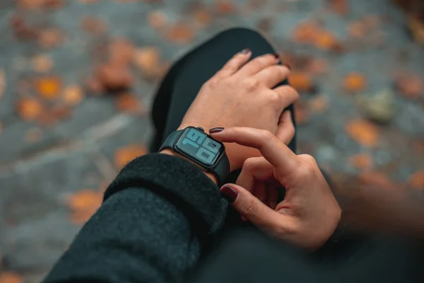 Photo of woman hands touching setting of digital smart watch in park. The time on the clock is 19-00.