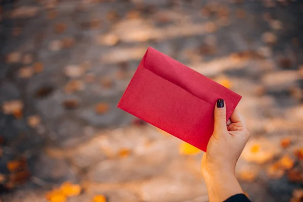 A woman opens a red envelope while sitting on a bench in a city park. Red envelope without inscriptions on a background of autumn foliage. High quality photo
