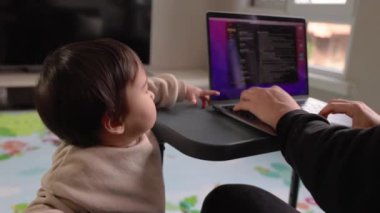 Bir adam evinde, kanepede dizüstü bilgisayarıyla çalışırken, bir yaşındaki çocuğu masaya tutunuyor. Yüksek kalite 4k görüntü