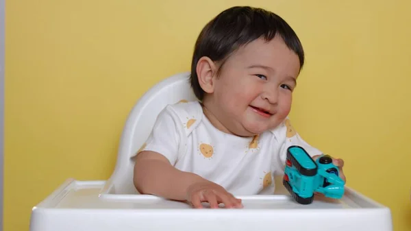 Aranyos Ázsiai Baba Etetőszéken Játékkal Mosolyog Sárga Háttér Kiváló Minőségű — Stock Fotó