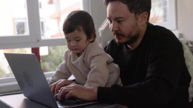 Dizüstü bilgisayarda çalışmaya çalışan genç bir adam oğlunu evde tutuyor. Uzaktan kumanda. Yüksek kalite 4k görüntü
