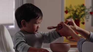 Asyalı bebek kendi başına yemek için annesinden bir kaşık almaya çalışıyor. Anne bebekten kaşık ve kase yiyecek alır. 4k görüntü