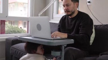 Bir adam evinde, kanepede dizüstü bilgisayarıyla çalışırken, bir yaşındaki çocuğu masaya tutunuyor. Yüksek kalite 4k görüntü