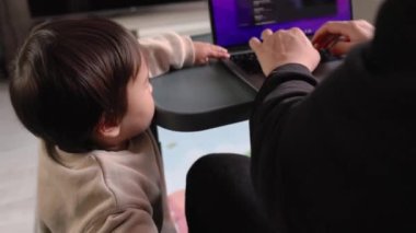 Bir adam evinde, kanepede dizüstü bilgisayarıyla çalışırken, bir yaşındaki çocuğu masaya tutunuyor. Yüksek kalite 4k görüntü