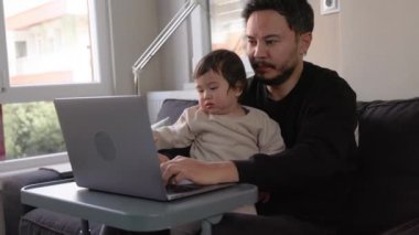 Dizüstü bilgisayarda çalışan genç bir adam oğlunu evde tutuyor. Uzaktan kumanda. Yüksek kalite 4k görüntü