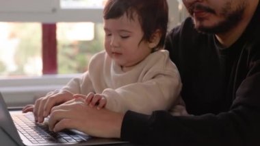 Dizüstü bilgisayarda çalışmaya çalışan genç bir adam oğlunu evde tutuyor. Uzaktan kumanda. Yüksek kalite 4k görüntü