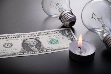 Electric lamp and candle on a dark background. Incandescent lamp, 1 dollar usa and a candle. Shutdown or blackout of electricity. High electricity prices. Saving electricity.