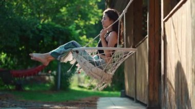 Yalınayak kız oturuyor ve sallanıyor örgü örülmüş hamak sandalyesine yaz günü bahçede boho tarzında, doğada dinleniyor.