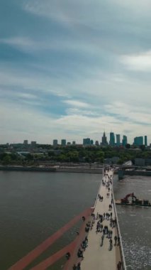 Günbatımında Vistula Nehri üzerinde yeni inşa edilen modern yaya köprüsü, şehir merkezindeki finans bölgesi ve Varşova şehrinin modern mimarisi, havadan görüş 4k. Yüksek kaliteli dikey görüntü