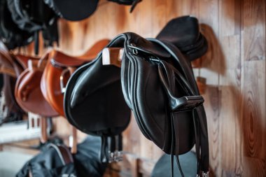 Ahır veya ahırdaki ahşap duvarda asılı siyah deri eyer, profesyonel binicilik ekipmanı. Yüksek kalite fotoğraf
