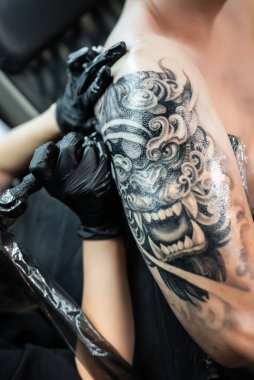 Profesyonel kadın sanatçı erkek elinde Japon şeytan maskesinin stil sahibi siyah dövmesini yapıyor kuaförde iğne ve makine ile, omuz vücut sanatı. Yüksek kalite fotoğraf