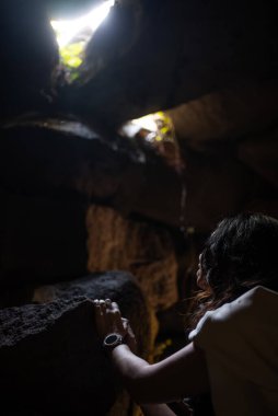 Kişi loş ışıklı mağaranın içinde keşfetmekle meşgul, kayalık yapıdaki doğal açıklıktan akan ışık ışınlarına dikkatle bakıyor.