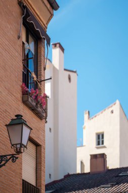 Çiçeklerle süslenmiş parlak balkon, canlı kentsel bir ortamda berrak mavi gökyüzünün altındaki yakınlardaki beyaz yapılarla çelişen tarihi bir tuğla binanın üzerinde yer alıyor. Yüksek kalite fotoğraf