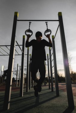 Spor salonunda yüzüklerini çalıştıran genç bir atletik gün batımında sokak spor salonunda barfiks çekiyor, vücut geliştirme ve dayanıklılık eğitimi kavramı. Yüksek kalite fotoğraf