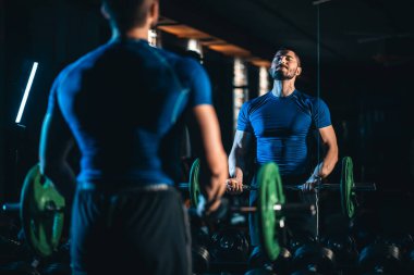 Erkek vücut geliştirici, spor salonunda pazılara halter kıvırarak ayakta duruyor, antreman sırasında ayna önünde halterle kol kaslarını çalıştırıyor. Yüksek kalite fotoğraf