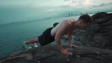 Erkek vücut geliştirici, gün batımında deniz kenarındaki kayalıklarda şınav çekiyor, erkek sporcu gün batımında okyanus uçurumunda şınav çekiyor. Yüksek kalite 4k görüntü