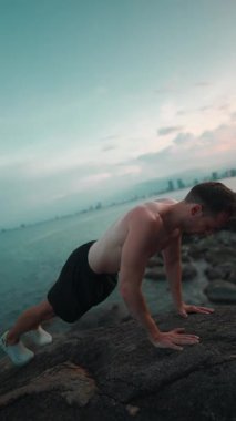 Erkek vücut geliştirici, gün batımında deniz kenarındaki kayalıklarda şınav çekiyor, erkek sporcu gün batımında okyanus uçurumunda şınav çekiyor. Yüksek kalite 4k görüntü, dikey video