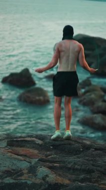 Güneş batarken kayalıklarda ip atlayan vücut geliştirici, gün batımında kardiyo egzersizi için deniz kıyısında ip atlama antrenmanı yapan sporcu. Yüksek kalite 4k görüntü, dikey video