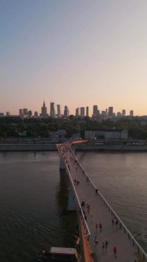 Günbatımında Vistula Nehri üzerinde yeni inşa edilen modern yaya köprüsü, şehir merkezindeki finans bölgesi ve Varşova şehrinin modern mimarisi, havadan görüş 4k. Yüksek kaliteli dikey görüntü