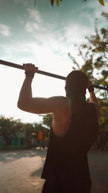Erkek vücut geliştirici gün batımında barfiks çekiyor, sporcu vücudunun üst kısmını güneş batımında barfiksle eğitiyor, 4k dikey video. Yüksek kaliteli görüntüler