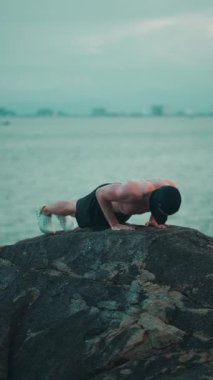 Erkek vücut geliştirici, gün batımında deniz kenarındaki kayalıklarda şınav çekiyor, erkek sporcu gün batımında okyanus uçurumunda şınav çekiyor. Yüksek kalite 4k görüntü, dikey video