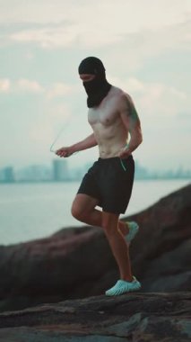 Güneş batarken kayalıklarda ip atlayan vücut geliştirici, gün batımında kardiyo egzersizi için deniz kıyısında ip atlama antrenmanı yapan sporcu. Yüksek kalite 4k görüntü, dikey video