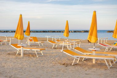 Güneşin batışında güzel bir sahil, sarı kum, mavi gökyüzü, deniz. Deniz sahilinde güneş pansiyonları ve kapalı şemsiyeler. İnsansız plaj. Tatil kavramı