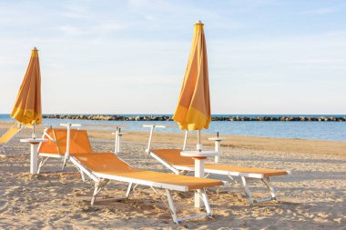 Güneşin batışında güzel bir sahil, sarı kum, mavi gökyüzü, deniz. Deniz sahilinde güneş pansiyonları ve kapalı şemsiyeler. İnsansız plaj. Tatil kavramı.