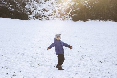 Güneşli bir kış gününde kolları açık, karla kaplı bir arazide duran mutlu çocuk. Kış çocukluğunu temsil eden kavram, soğuk mevsimlerde boş zaman aktiviteleri ve aile eğlencesi.