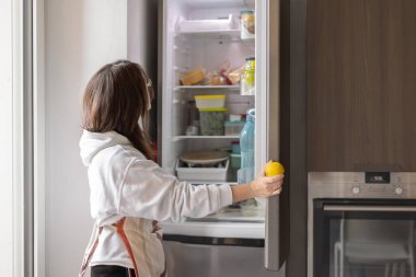 Genç bir kadın modern mutfakta buzdolabı açıyor. Elinde limon, sağlıklı yiyecek konsepti var..