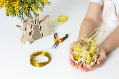 El yapımı altın telden ve sarı kurdeleden yapılmış Paskalya yumurtalarını tutan kadınların yakın plan fotoğrafları. Masada DIY aletleri ve mimoza var..