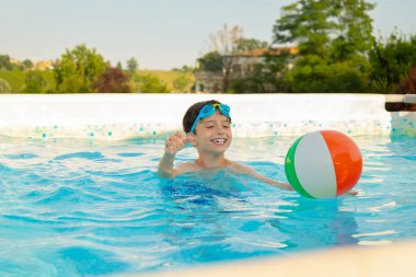 Gözlük takan gülümseyen çocuk yaz tatilinde yüzme havuzunda şişme bir topla oynuyor, su oyunlarıyla eğleniyor ve güneşli günün tadını çıkarıyor.