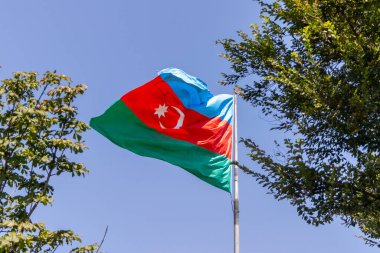 Bakü, Azerbaycan-02 Ağustos 2024: Azerbaijan 'ın ulusal bayrağı açık mavi gökyüzüne karşı gururla sallanıyor, yemyeşil ağaçlarla çevrili, ulusal gurur ve kimliği simgeliyor