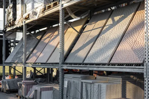 Bir depo veya perakende ortamda metal raflarda çeşitli renkler, dokular ve desenler gösteren geniş formatlı fayanslar düzgünce düzenlenmiştir.