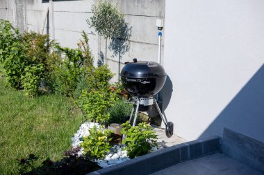 Piacenza, İtalya - 11 Mayıs 2025: Siyah çaydanlık ızgara Weber yeşil çimenli bir bahçede tekerlekler üzerinde duruyor, çeşitli bitkiler güneşli bir yaz günü barbekü partisine hazır bir ev duvarının yanında