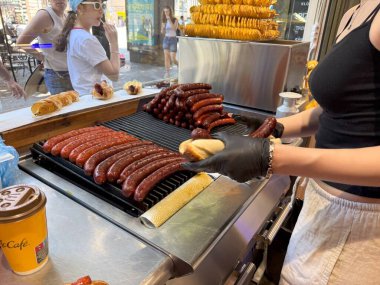 Prag, Çek Cumhuriyeti - 22 Haziran 2025: Siyah eldivenli sokak yemekleri satıcısı taze ızgara sosisi açık büfede yumuşak bir çöreğe yerleştiriyor, çıtır patates cipsi sergiliyor ve cezbedici.