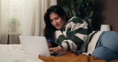 Yatak odasında uzanan güzel bir kadın egzotik ülkelere seyahat satın alıyor, online seyahat acentası kullanıyor, rezerve edilebilir otel kullanıyor, tatilini kredi kartıyla ödüyor..