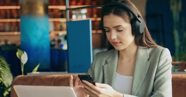 穿着时髦西服 戴着无线耳机 漂亮可爱的年轻女子坐在餐馆里 通过手机和笔记本电脑在网上工作 自由职业者时间概念工作流程 — 图库视频影像