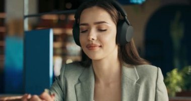 Close up shot selective focus on young buisness lady woman sitting at table wearing wireless headphones listens to music while eating dessert at cafe having break from work. Dream job.